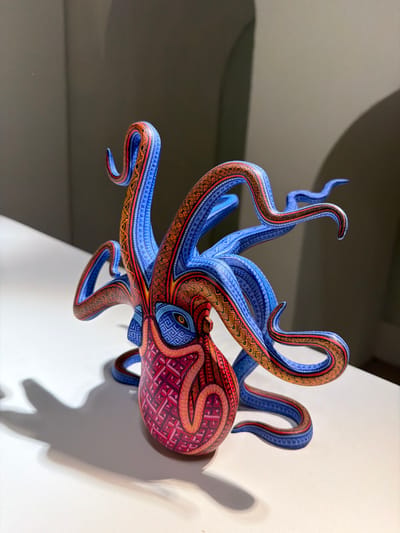 A vibrant, detailed wooden octopus sculpture clings to a white wall. Its body is adorned with a red and pink geometric pattern, transitioning to blue and orange on the tentacles. The tentacles, painted in intricate patterns, twist and curl, creating dynamic shapes. A shadow of the octopus is visible on the wall, and a soft, diffused light illuminates the sculpture. The backdrop consists of plain walls, highlighting the craftsmanship and artistry of the piece.