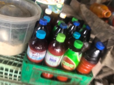 The image shows a close-up of a stack of plastic bottles of various colors. Most of the bottles are dark in color and have a blue cap. There are also two red bottles with white labels, a green bottle with a white label, and a white plastic container with a green lid. The bottles are arranged in a semi-circle shape. The background is a blur of various objects, but it is possible that the location of the image is a small shop or market in Umudike, Nigeria. The coordinates provided correspond to a location in Umudike. The image is likely taken in a grocery store or market, with a focus on the bottles of drink and snacks, giving the impression of the scene being in Umudike, Nigeria. There are no people in the image.