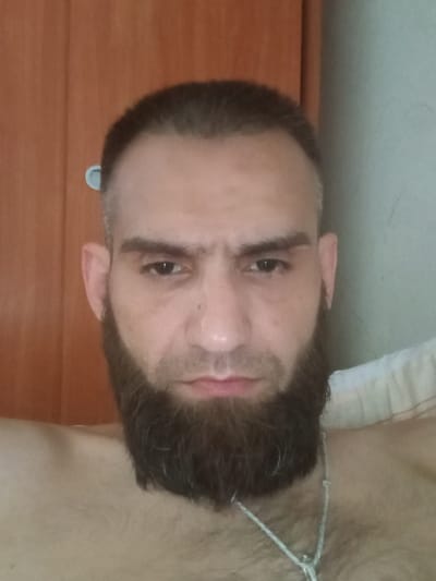The image shows a close-up, head-and-shoulders portrait of a man with a full beard and short hair. He is looking directly at the camera with a neutral expression. The background consists of a wooden surface on the left and a plain, light-colored wall on the right. The lighting appears to be natural, coming from the front. There are no discernible landmarks or features that would suggest a specific location within the city of Rezekne, Latvia. The man is wearing a silver necklace.