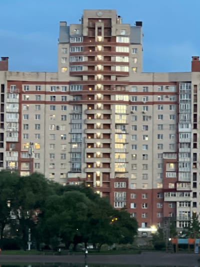 The image shows a tall, multi-story building with a facade of light beige and reddish-brown brick. The building has numerous windows, some of which are illuminated, suggesting it is either evening or night. The architecture appears to be residential, with balconies and varied window styles. To the right of the building, there is a dense cluster of dark green trees, partially obscuring the lower sections of the building and extending upwards. The sky above is a muted blue, indicating dusk or a cloudy day. A dark gray pole or fence runs vertically along the right edge of the frame, and there is a hint of other structures with colorful elements in the far background, suggesting an urban or suburban environment. No people are visible, and no text is present. The overall impression is a quiet, perhaps residential scene at the transition from day to night.