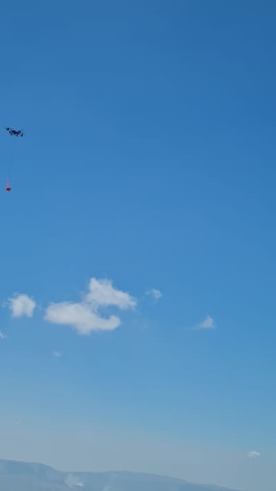 The image shows a clear blue sky with a few scattered white clouds. In the upper left quadrant, a black drone is visible, hovering in the air. A small, red object, possibly a bucket or container, is suspended from the drone by a thin line. The drone and its payload are positioned against the vast expanse of the sky. In the distance, at the bottom of the image, a hazy, mountainous landscape is discernible, with faint plumes of smoke rising from the terrain, suggesting a potential wildfire or controlled burn in the L'Aquila, Italy area. The overall scene conveys a sense of aerial observation or intervention.
