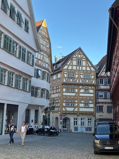 This image depicts a sunlit cobblestone square in Esslingen am Neckar, Germany, with a couple walking hand-in-hand in the foreground. They are positioned on the left side of the frame, with the man on the left wearing a patterned short-sleeved shirt and dark jeans, and the woman on the right in a white tank top and linen pants. Behind them, a white building with numerous green shutters lines the left side of the square, while to the right, traditional timber-framed buildings with white infill and exposed wooden beams dominate the scene. Several outdoor tables and umbrellas suggest a cafe or restaurant setting in the middle ground. A black Mercedes-Benz van is parked on the right side of the square, facing the viewer. The architecture suggests a historic town center.