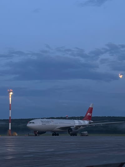 This image captures a Nordwind Airlines airplane parked on the tarmac at Emelyanovo Airport in Russia. The scene is set during twilight, as indicated by the dusky blue sky with scattered clouds and the faint illumination of the moon. The airplane, predominantly white with red accents and the Nordwind logo, is the central object. The tail fin displays a prominent red 'N'. The aircraft's engines are visible, as are its landing gear.
In the foreground, a red and white striped barrier with lights is positioned, likely a runway or taxiway marker. The tarmac itself appears wet, suggesting recent rain or damp conditions. Beyond the aircraft, a line of trees forms a dark silhouette against the horizon, hinting at a natural environment surrounding the airport. The overall mood is calm and serene, typical of an airport at the end of the day. No people are clearly visible in the frame. Visible text includes "Nordwind" and "Airlines" on the side of the aircraft.