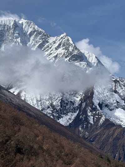This image presents a majestic high-altitude mountain landscape, characteristic of the Khumjung region in Nepal. The dominant subject is a towering, snow-capped mountain range, occupying the left and central parts of the frame. The peaks are rugged and covered extensively in pristine white snow and ice, with darker, exposed rock faces visible throughout. Swirling clouds and mist partially obscure the mid-slopes and peaks of the mountains, creating a dynamic and atmospheric effect.
To the right of the formidable snowy peaks, a steep slope or ridge is visible, covered in dry, brown vegetation, likely bare trees or shrubs, offering a contrasting texture and color to the snow and rock. The upper left portion of the image reveals a clear, vibrant blue sky with minimal cloud cover.
The scene captures a static natural environment; no specific activity or event is taking place, and no people or animals are visible. It is daytime, with bright, natural light illuminating the landscape. The weather appears cold given the snow, but the clear sky suggests good visibility above the immediate mountain clouds. The overall impression is one of immense scale, tranquility, and the raw beauty of the Himalayan wilderness. No text is visible in the image.