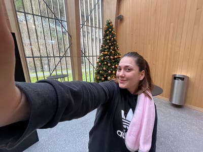 An adult female is depicted taking a selfie indoors, located in Erstfeld, Switzerland. The woman is positioned centrally, extending her right arm to hold a device (presumably a smartphone) for the photograph. She wears a black hooded sweatshirt with an Adidas logo and has a light pink towel draped over her left shoulder. Her hair is pulled back into a ponytail, and she exhibits a subtle smile.
In the background, to the right of the woman, stands a Christmas tree adorned with numerous gold-colored spherical ornaments. Adjacent to this, a light wood-paneled wall hosts a cylindrical stainless steel trash receptacle with a dark opening. Two small, dark, circular tables are visible; one is partially obscured behind the woman, and another is positioned outside the prominent, multi-paned windows. These large windows, featuring dark framing and diagonal cross-braces, dominate the left and background portions of the scene, offering a view of an outdoor environment. This outdoor area includes barren trees, green ground cover, a paved path, and a distant rocky hillside. The indoor flooring is a grey, textured material, possibly tile.