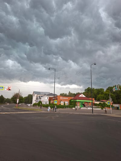 This image depicts a street scene in Murom, Russia, under a dark, stormy sky. The foreground is dominated by a wide asphalt intersection with painted white pedestrian crossings. In the midground, a street curves to the left, with parked cars lining the right side. Several buildings are visible: a light-colored building with arched windows on the left, a red brick building with a green facade and a large "5" logo (indicating a Pyaterochka supermarket) in the center, and other structures partially obscured by trees.
The sky is filled with heavy, dark gray clouds, suggesting an impending storm. The lighting is dim, casting a somber mood over the scene.
Several individuals are present:
* Two pedestrians are walking across the street from left to right.
* A woman in a light-colored dress is walking with a young child in a blue dress.
* An older man is riding a bicycle towards the right side of the frame.
* A person, possibly male, is standing on the sidewalk to the right of the Pyaterochka store.
* A group of people are gathered near the entrance of the Pyaterochka store.
* Several individuals are seen walking on the sidewalk in front of the light-colored building on the left.
Traffic signals are visible, including one with a red light and another with a green light. Pedestrian crossing signs with blue squares and white figures are posted at intersections. Streetlights with single black poles and lamps are positioned on either side of the road. Trees line the sides of the street, with lush green foliage.