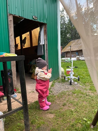 An outdoor, eye-level, medium shot captures a young child standing on a gravelly patch of ground in front of a green corrugated metal structure. The child, wearing a grey dotted hat, a patterned jacket, and pink overalls with matching pink boots, faces away from the camera and appears to be clapping their hands. A white child's tricycle with a red bucket is visible to the right of the child, on a grassy area. In the background, beyond the tricycle, is a wooden structure with a corrugated roof, and further beyond that, trees and a cloudy sky. To the left of the child, a black metal structure supports a black speaker with red accents. The ground is a mixture of gravel and grass, with some areas appearing damp. The overall scene suggests a rural or semi-rural outdoor setting. The location is identified as Azinsky, Russia.