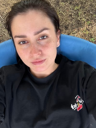 A close-up, overhead shot shows a woman lying down outdoors, looking up at the camera. She has dark hair, light skin with freckles, and is wearing a black t-shirt. She is partially reclined on a blue cushioned surface, which is set against a backdrop of dry grass and dirt. The image is sharp and well-lit, with the woman's face as the primary focus. The scene appears to be in a natural outdoor setting, possibly a park or backyard. There are no other people or animals visible in the frame. The overall impression is one of casual relaxation in an outdoor environment.