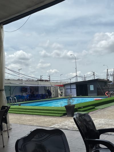 The image shows an outdoor swimming pool area in what appears to be an urban setting, likely in Port Harcourt, Nigeria. The pool has blue water, and its perimeter is lined with artificial green turf. Along one side of the pool, there is a structure with blue chairs and a fence topped with barbed wire. Utility poles and wires are visible above the structure. A life preserver is mounted on the wall.
The sky is cloudy. In the foreground, there are black chairs. The photograph is taken from a low angle, and the camera is positioned close to the chairs. The composition is oriented vertically.