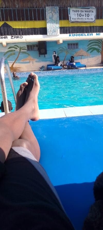 The image captures a first-person view of a poolside scene on a bright, sunny day, conveying a sense of relaxation. In the immediate foreground, a person's bare, dark-skinned legs are visible, crossed at the ankles, resting on a bright blue surface, likely a lounge chair or mat. The person is wearing dark shorts with a light-colored waistband.

Beyond the legs, the vibrant blue water of a swimming pool fills the midground, showing ripples and reflections from the sunlight. The pool has a white or light-colored tiled edge with a border of blue patterned tiles. A silver metal handrail for pool entry is visible on the left side of the pool. Two individuals are partially visible in the water: one's head and shoulders are on the far left, and another person's dark head is barely peeking out from the bottom left edge of the frame.

In the background, a building with a brightly painted mural forms the backdrop to the pool. The mural depicts a serene beach scene with two prominent palm trees with green fronds, two straw umbrellas, and sand-colored ground. Two painted figures are also part of the mural, one appearing to wear a white dress. In front of this mural, two people are seated on blue lounge chairs by the poolside, both wearing dark clothing, appearing to relax or converse.

Above the mural, the building features a corrugated metal roof section painted in distinctive yellow and black horizontal stripes. Attached to this structure are several items: a rectangular white relief sculpture or artwork with a light blue, abstract figure is mounted on the left. A black speaker is attached to the wall below the roofline, to the left of the painted figures in the mural. Windows are also visible on the wall above the mural.

Several pieces of text are visible on the building:
- On the left side of the building, above the mural, partially cropped, the words "AHA ZAKO" are visible, possibly part of a larger phrase like "RAHA ZAKO".
- A white banner hanging from the roof structure on the right side displays "TUZU ZA WASTA", followed by a "10+10" graphic with two small stick figures, and below that, "Kwa Ufasaha na Mantiki".
- Below this banner, on the wall, the phrase "KUOGELEA NI" is visible, also partially cropped.

The strong sunlight and clear reflections on the water indicate it is daytime, likely a warm and pleasant day. The overall atmosphere suggests leisure and enjoyment by the pool.