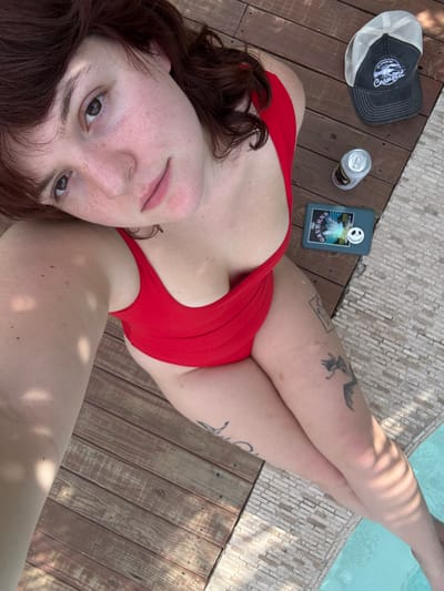 A woman in a red swimsuit poses near a pool. She is lying on a wooden deck next to white tiled coping. A baseball cap, a can, and a phone rest nearby. The woman has fair skin and reddish-brown hair. She has tattoos on her legs. The pool water is visible. The photo is taken from a high angle, showing the woman's face and body.