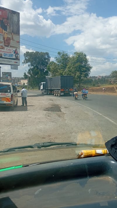 A truck transports a dark-colored container on a sunny day. Two motorbikes follow closely. A man stands beside a minivan labeled "GRACE GIL-RISH Shuttle." The road is wide and dusty. A billboard is visible in the background, advertising drinks. The image is taken from inside a vehicle, with the dashboard and partial windshield visible. The sky is blue with scattered white clouds.