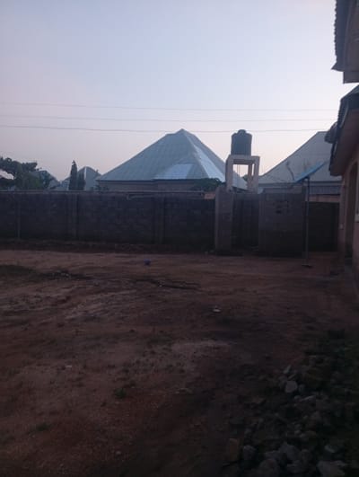 The image depicts an outdoor scene in Zanzi, Nigeria, characterized by a low wall constructed from dark, rectangular bricks. Beyond the wall, several houses are visible. One house features a distinctive pyramid-shaped roof made of corrugated metal. Another structure has a water tank mounted on a concrete support. The ground in the foreground is uneven and appears to be dirt or soil, with scattered rocks and some sparse vegetation. The sky is a pale, hazy blue, suggesting it may be early morning or late afternoon. There are no people or animals present in the image.