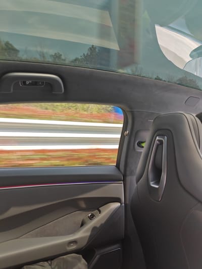 The scene is an interior view from a moving vehicle in Jurong, China, observed from a rear passenger's perspective. A panoramic glass roof reveals a blurred overhead sky and indistinct treetops. Through the side window, a landscape in rapid motion is depicted by horizontal streaks of white and gray, consistent with a road barrier or guardrail, interspersed with blurred orange, red, and green tones of foliage. The vehicle's interior on the left includes a gray door panel with an integrated armrest, a window control switch, a door handle, and an ambient light strip displaying distinct red and blue/purple illumination. An overhead grab handle is positioned above the door. In the lower left footwell, a dark, crumpled object, possibly clothing or a bag, is visible. On the right, the upper portion of a black, bolstered passenger seat backrest is visible, featuring a metallic-silver colored accent or handle. The adjacent B-pillar is upholstered in gray material and bears an "AIRBAG" label; a bright green object is partially recessed within an opening on an interior panel near the B-pillar.