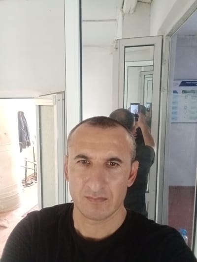 The image is a selfie taken by a man in a black t-shirt, positioned in the foreground. He has short dark hair and is looking directly at the camera. In the background, a reflection in a mirrored door shows the man taking the photo with a smartphone. Further reflections in a series of doors reveal a hallway with a bulletin board on the right wall, displaying text and diagrams. On the left side of the background, a large white cylindrical object is visible, along with a metal rack with a dark garment hanging from it. The environment appears to be an indoor space, possibly an office or a public building, with white walls and doors. No specific identifiable landmarks indicating the city of Karagana, Uzbekistan, are present in the image.
