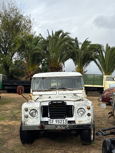 The image depicts a weathered, white Land Rover parked on a grassy terrain near a palm tree. The license plate reads "CF 98213". The vehicle shows signs of rust and disrepair. In the background, parts of other trucks are visible. The sky is overcast. The location is indicated to be Kraaifontein, South Africa.