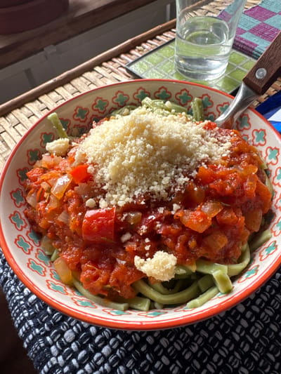 A bowl of green pasta, topped with a chunky red tomato sauce and a mound of grated Parmesan cheese. The sauce contains diced tomatoes, onions, and possibly other vegetables. A fork rests in the bowl. The bowl sits on a wicker tray beside a glass of water on a green and purple patterned mat. A black and white patterned fabric is visible on the right side of the frame.