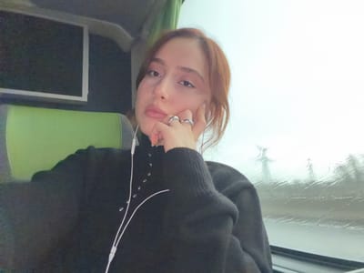 A female individual is seated within a moving vehicle, likely a bus or coach, during daytime overcast or rainy conditions. She is positioned in the foreground, facing the camera directly, capturing a self-portrait. She has reddish-brown hair and fair skin. Her left hand, adorned with a silver-colored ring on the ring finger, is raised with the index finger and thumb supporting her chin. She wears a dark, long-sleeved garment with a high collar and multiple small, metallic studs or buttons arranged vertically on the left chest area. White wired earphones are inserted into her ears, with the wires visible descending along the front of her top. Behind her left shoulder, a distinctive green and grey padded headrest or upper seat portion is visible. To her upper left, a rectangular screen with a dark display is mounted on the vehicle's interior wall. To her right, a window reveals an exterior scene characterized by streaks and droplets of water on the glass, indicative of rain. Through the wet window, indistinct, dark silhouettes, resembling power line towers, are visible against a bright, uniformly overcast sky. The image does not contain specific visual information to independently verify the location as Moineville, France.