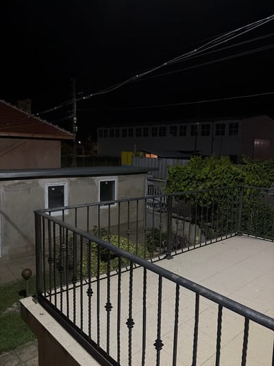 The image captures an outdoor scene at night, likely in the city of Dolni Dabnik, Bulgaria, focusing on a balcony area. The foreground features a light-colored tiled floor on a balcony, bordered by a black wrought-iron railing with decorative twisted spindles. To the left, a dark-colored metal handrail runs along the top of the railing.
Beyond the balcony, a low wall or structure is visible, constructed with rough, textured concrete. Two windows with white frames are set into this wall; their panes are dark, indicating that they are either closed, opaque, or unlit from within. A dark, spherical garden ornament on a thin pole stands in the grassy area to the left of this wall.
Further in the background, a red-tiled roof of a building is partially visible on the left. To the right of the wall with the windows, a dense green bush or tree thrives. Behind this, a dark, metal fence or gate is partially visible, with some indistinct structures behind it. The farther background is dominated by a long, low building with a dark facade and numerous evenly spaced rectangular windows that appear dark. Overhead, multiple power lines crisscross the dark night sky, supported by a utility pole. The overall atmosphere is dark and still.