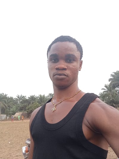 A young man is centered in the image, wearing a black tank top and a gold necklace with a cross pendant. He has short, dark hair and a serious expression. The background shows a grassy field with palm trees and some indistinct figures in the distance. The sky is overcast, suggesting a cloudy day. A portion of what appears to be a vehicle is visible in the lower left corner of the frame.