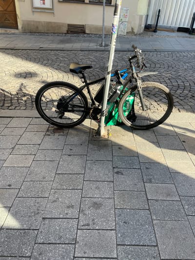 A black electric bicycle is parked on a cobblestone street next to a lamppost. The bicycle has a green bag attached to its frame. The street is lined with stone buildings, and the sun is casting shadows on the ground. In the background, there is a construction site with a metal fence and scaffolding. The location is identified as the area around Masaryk University in Brno, Czechia.