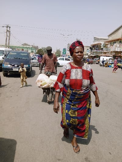 An outdoor scene depicts a paved street in Mumbwa, Zambia, under a bright, hazy sky. In the immediate foreground, a woman of apparent middle age walks towards the viewer's right, her body angled slightly left. She wears a red, black, and white patterned headwrap, a long-sleeved top with a similar red and white geometric pattern, and a long skirt in blue, red, and yellow patterns. She has dark-toned skin and wears dark flip-flops. Her gaze is directed generally forward and slightly to the left.
Behind her, slightly to the left, a man with dark-toned skin, wearing a patterned short-sleeved shirt and dark trousers, pushes a two-wheeled cart laden with multiple white sacks or bundles of produce, possibly peanuts or similar items. The cart has a wooden frame.
Further back and to the left of the man, a young child in a light-colored, long-sleeved garment stands near a parked dark blue Volkswagen minivan. The child appears to be looking towards the man and cart.
The street surface is asphalt, with faint tire tracks visible. In the background, numerous other vehicles, including cars and what appear to be motorcycles or tuk-tuks, are visible. On the right side of the street, multi-story commercial buildings line the road, some with open storefronts and awnings, suggesting a market or business district. Utility poles with overhead wires are present on both sides of the street. The overall impression is a bustling urban environment during daylight hours.