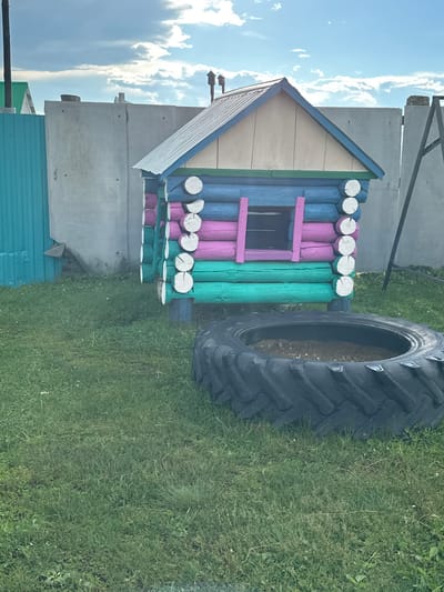 The image shows a brightly colored, miniature log cabin in a grassy yard. The cabin has a metal roof and is painted with blue, pink, and green logs. A large tractor tire filled with dirt sits in front of the cabin. In the background, a grey concrete wall and a turquoise fence are visible. The sky is cloudy. The location is identified as Urazovo, Russia.