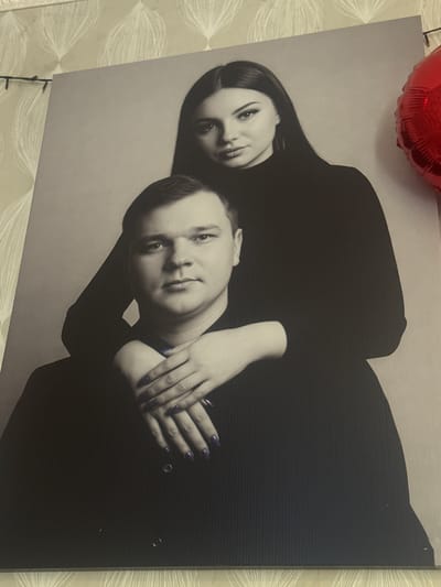 A black and white studio portrait features a man and a woman. The man is in the foreground, looking directly at the camera with a neutral expression. The woman is positioned behind him, with her arms wrapped around his neck and her hands resting on his chest. She is also looking towards the camera with a slight smile. Both individuals are wearing dark, long-sleeved tops. The background is plain and light-colored. The portrait is mounted on a surface that has a textured pattern, possibly wallpaper, visible at the top and bottom of the image. A red, heart-shaped balloon is partially visible in the upper right corner of the frame. The location of this photograph is not specified as being in the city of Razvedchik, Russia.