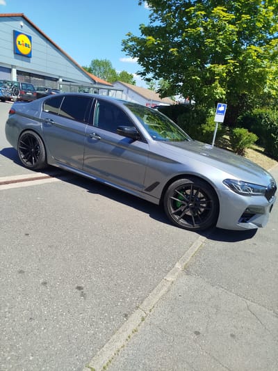 A grey BMW 5 Series sedan is parked in a parking lot in front of a Lidl supermarket. The car is viewed from the side, with the front of the car facing towards the right of the image. The car has tinted windows and black rims with green brake calipers. The Lidl store has a large yellow and blue logo on its facade. The parking lot is paved and there are trees and bushes in the background. The sky is blue and clear. The location is Kitzingen, Germany.