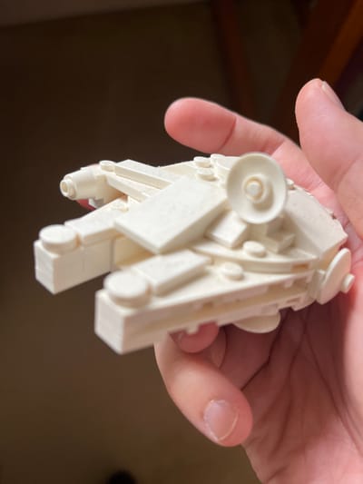 A close-up, slightly angled photograph captures a person's hand holding a small, intricate model constructed from white building blocks. The model, resembling a miniature spaceship or fictional vehicle, is the central focus, showcasing various studs, plates, sloped pieces, and circular dish-like elements, characteristic of a LEGO-style build. The hand, with light skin and visible fingernails on the thumb and index finger, gently holds the model. The background is softly blurred and dark, suggesting an indoor environment, with indistinct shapes possibly indicating furniture or a wall. The lighting is even, illuminating the white model clearly. The image captures the act of holding and presenting the detailed construction. There is no visible text or specific location cues within the image to identify Middle Park, Australia, nor are any emotions discernible from the partial view of the hand.