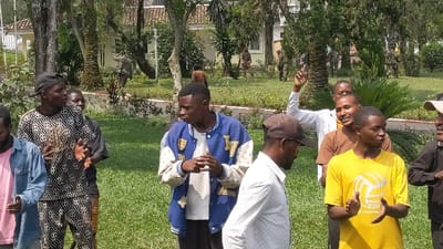 A group of young men stand on a grassy lawn, some clapping. One man wears a blue and white varsity jacket, while another has on a yellow t-shirt with a volleyball graphic. Some men are dressed in casual wear. In the background, other figures walk on the lawn, near trees and buildings. Palm trees and other green foliage are visible. The scene appears to be outdoors during the daytime.
