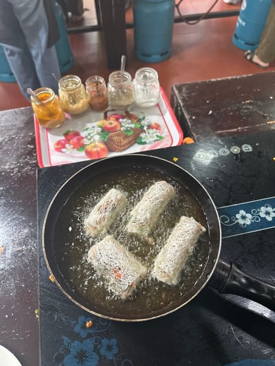 A frying pan on a stovetop contains four rolls being deep-fried in oil. Next to the stovetop is a tray with an apple design holding a row of spice jars with spoons. In the background, there are blue propane tanks. The scene appears to be a cooking setup.