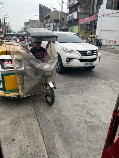 The image shows a street scene in Dagupan, Philippines. In the foreground, a tricycle, a common mode of transport in the Philippines, is partially visible. A driver wearing sunglasses and a black t-shirt is seated in the tricycle, which is covered with a transparent plastic sheet. The tricycle has a yellow and green color scheme and displays the number "33" on its side. In the background, a white SUV is parked on the street. The SUV has a license plate that reads "IAB 8630". Buildings and power lines line the street, indicating an urban environment. The sky is overcast, suggesting cloudy weather. The scene appears to be a typical street in a Philippine city, with vehicles and people engaged in daily activities.
