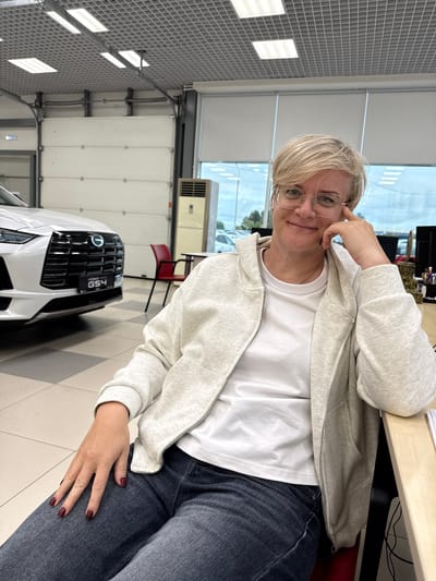 A medium shot, taken from a slightly low angle, shows a woman with short blonde hair and glasses sitting in a brightly lit indoor space, likely a car dealership in Yemelyanovo, Russia. She is wearing a light-colored hooded jacket over a white t-shirt and dark denim jeans. Her right hand is resting on her cheek, and she is looking directly at the camera with a slight smile. Her fingernails are painted a dark red.
In the background, a white car with a prominent black grille and "GS4" badging is partially visible to the left. A red chair is positioned behind the car, and a tall, beige air conditioning unit stands to the right of the car. The environment appears to be a showroom with a tiled floor and a high ceiling with fluorescent lighting and a grid structure. Large garage doors are visible in the background. A wooden desk is present to the right of the woman, with some papers and office supplies on it. The overall lighting suggests daytime.