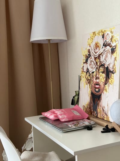 The image shows a white desk against a wall. On the desk, there is a laptop, two pink packets, a framed painting of a woman with flowers on her head, and a green bottle with reeds. To the left of the desk, there is a tan curtain and a floor lamp with a white shade. A white chair is partially visible to the left of the desk. The scene is set indoors, likely in a bedroom or living area, but there is no specific indication of the city of Semiluki, Russia.