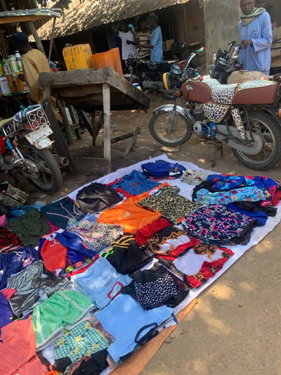 The image depicts an outdoor market scene in Nigeria, likely in the morning or late afternoon given the dappled sunlight and shadows. The foreground features a spread of colorful clothing laid out on a white cloth, interspersed with more items on the ground. The clothing includes a variety of patterns and colors, such as floral prints, leopard print, polka dots, and solid bright hues like orange and green.
Behind the clothing display, several motorcycles are parked. One motorcycle, a brown and tan model with a distinctive patterned seat, is prominently featured. The motorcycles suggest a mode of transportation and potentially part of the market's setup.
In the background, there are structures that appear to be market stalls or simple buildings, constructed with corrugated metal roofing and wooden beams. Several individuals are visible, some appearing to be vendors or customers. One person in the upper left is wearing a light blue robe and head covering. Another person is leaning over a motorcycle in the middle left. Further back, other figures are less distinct.
On the left side of the image, several large yellow and orange plastic containers are stacked, possibly used for storage or as part of the market's goods. Some bottles and other items are also visible on a table-like structure. The overall scene conveys a bustling, informal market environment. No specific text is clearly legible in the image.