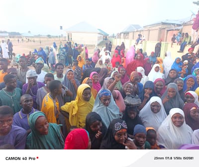 A daytime wide-angle shot depicts a large group of people gathered in an open area in Wase, Nigeria. The crowd, consisting of adults and children, are densely packed and face towards the camera. The majority of the women wear colorful headscarves, while many men wear casual attire. The scene includes an open field with some scattered people in the background, a building, and a wall. The image details indicate the photo was taken with a CAMON 40 Pro camera, with settings 23mm f/1.8 1/50s ISO581.