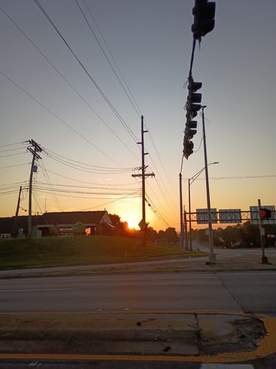 This is a vertically oriented, medium-shot, eye-level photograph of a street scene at dusk. The sky is a gradient of pale blue at the top transitioning to a bright orange-yellow near the horizon, indicating either sunrise or sunset. Silhouetted against the sky are traffic lights and utility poles with numerous wires. In the background, trees line the horizon, and a few buildings are visible, one with a green sign that appears to read "RUSTIC GRILLE." A road runs diagonally through the scene, with yellow painted lines and a cracked asphalt surface. The overall mood is peaceful and serene, with the warm light of dusk casting long shadows and bathing the scene in a soft glow. There are no people visible in the image.