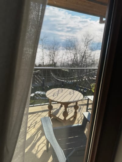 The image depicts a view looking outward from an interior space, through a mesh screen covering a portal, onto a sunlit balcony in Sighnaghi, Georgia. A light-colored, textured fabric curtain partially obscures the left side of the frame, while a dark wooden frame borders the right.
The balcony features a light-toned floor, likely concrete or tile, strongly illuminated by sunlight which casts distinct diagonal shadows from the balcony railing and furniture. A round, light-colored wooden table with a central pedestal base stands centrally on the balcony. To its left, a light-toned, possibly grey, outdoor chair is partially visible. To the right of the table, another darker chair is partially visible. The balcony is bordered by a dark metal railing with a decorative, repeating pattern.
Beyond the balcony, the background shows a partially cloudy sky with prominent white clouds and patches of blue. Numerous bare deciduous tree branches are visible against the sky, suggesting a dormant season. Some faint green foliage is present towards the lower right. The overall scene indicates an elevated perspective overlooking a landscape, consistent with the city of Sighnaghi. No individuals or specific city structures are visible beyond the immediate balcony and natural elements.