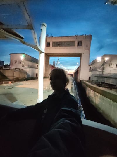 A person, with light-colored hair and wearing a dark jacket, is standing on what appears to be a boat or pier at dusk. The person is facing away from the camera and looking towards a large, concrete structure that spans across a waterway. The structure has multiple windows and the number "8" is visible on its side. In the background, there are two light-colored buildings with windows, and streetlights are illuminated. The sky is a deep blue, indicating the time of day is either early evening or pre-dawn. The scene suggests an industrial or port area, possibly a lock or dam. The location is identified as Sheksna, Russia.