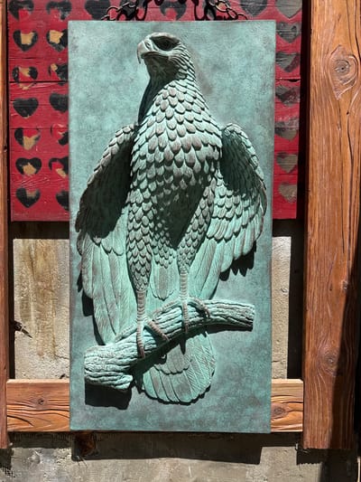 An outdoor, eye-level, medium shot captures a relief sculpture of a raptor perched on a branch, mounted on a wall. The sculpture is rendered in a verdigris patina, accentuating the detailed feathers and sharp talons. The background is a rustic wooden wall with a red panel featuring a pattern of black and gold hearts. To the right of the sculpture, a large, roughly hewn wooden beam is visible. The sculpture is likely located in Kolomyia, Ukraine, given the context. The scene is lit by natural sunlight, casting distinct shadows that highlight the three-dimensional quality of the artwork.