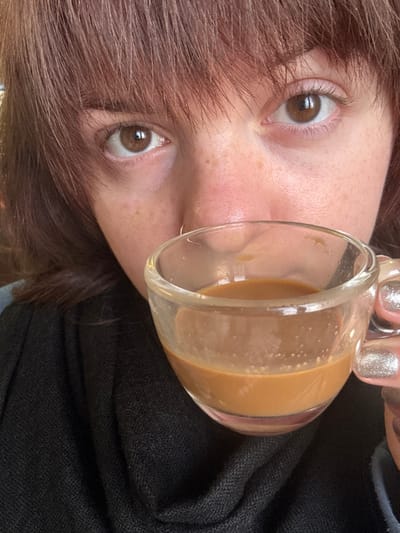 A person with light skin, brown hair, and brown eyes holds a clear glass mug containing a light brown liquid. The mug is positioned near their face, partially obscuring the view. The person wears silver nail polish. A black garment is visible on the side. The image is rotated, displaying the subject at an angle. The person has a nose ring.
