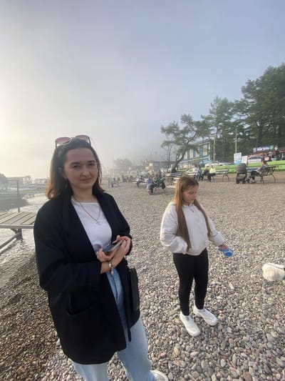 In Listvyanka, Russia, two females are present on a pebbled shoreline. The woman in the foreground, on the left, is wearing a black blazer over a white shirt and light-colored jeans. She has sunglasses perched on her head and is holding a mobile phone in her right hand. A young girl, on the right, is dressed in a cream-colored hooded jacket and black leggings. She is holding a blue leash. The background reveals a misty, overcast sky, with trees and buildings situated further inland. Several other people are visible in the background, some sitting on benches. The water of a lake is visible on the far left of the frame.