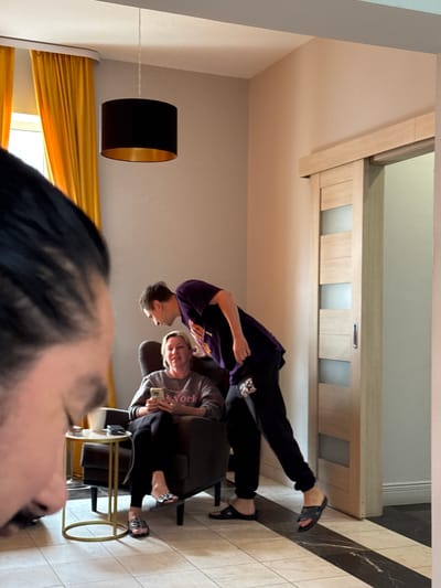 A woman is sitting in a chair holding a phone. A man is leaning over her, looking at her phone. The room has yellow curtains and a sliding door. The floor is tiled with black and white squares. The scene appears to be indoors, in an apartment or house. The location is identified as Ptichnoye, Russia.