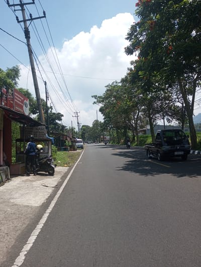 The image depicts a road scene on a sunny day. On the left side of the road, there's a roadside eatery or stall. A person in a blue shirt stands near a motorcycle parked beside the stall. The stall has a sign with the words "WARUNG NASI" visible. Power lines and poles run along the left side of the road. The road itself is paved and gray, with a white line marking the edge. A black pickup truck is parked on the right side of the road, near trees with reddish-orange flowers. Two motorcycles are seen further down the road. Other vehicles and a building are visible in the distance. The sky is blue with scattered white clouds. There is no indication of this being the city of Nawng Hkar, Myanmar.