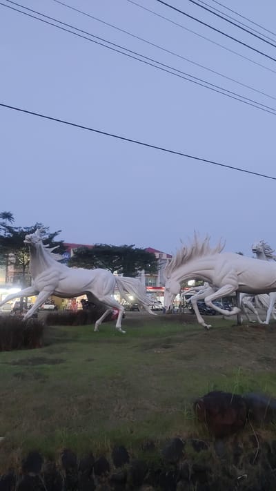 In the city of Pusakarakyat, Indonesia, a sculpture of white horses running is displayed on a grassy median. The sculptures are in various dynamic poses, some appearing to gallop. In the background, buildings, trees, and a few vehicles can be seen. The sky is a pale, overcast blue, and power lines are visible stretching across the frame. The foreground features a manicured lawn with some dark rocks at the bottom edge. The overall impression is of a public art installation in an urban setting.