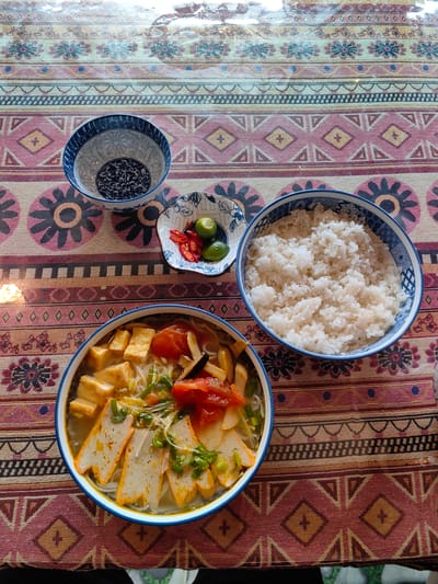 The image shows a table setting for a meal. The setting consists of a bowl of noodle soup with tofu and tomatoes, a bowl of rice, a bowl with dark-colored seeds or beans, and a small dish containing sliced red chilies and lime wedges. The bowls are blue and white patterned. The meal is set on a patterned tablecloth.
The location is likely Thành phố Hà Giang, Vietnam, based on the user’s prompt.
