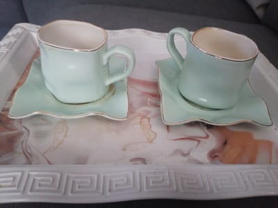 Two pale blue ceramic coffee cups with gold rims rest on matching saucers, which are placed on a rectangular white tray with a decorative border. The tray has a marbled pattern of pink and brown hues. Both cups have visible stains on the inside rim. The scene appears to be indoors, with a grey upholstered surface visible in the background. The image was taken at a slight angle, looking down at the objects. There is no indication of the city of Silifke, Turkey, or any specific location details.