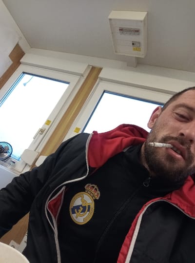 The image displays an indoor scene in the city of Drugan, Bulgaria, featuring one adult male. The male subject, possessing a beard and dark hair, is positioned in a low-angle view, facing the camera. He is wearing a black tracksuit jacket with a prominent yellow and white Real Madrid crest logo on the left chest, featuring a red hood and red internal lining. A lit cigarette is held between his parted lips.

In the background, two large, light-colored framed windows dominate the upper left and central portions of the frame. The external view through the windows is uniformly bright white, lacking discernible outdoor features. Multiple small, yellow rectangular stickers are affixed to the internal window frames. A small, black electric fan is situated on a surface, possibly a windowsill, beneath the left window. Above the windows, a rectangular, off-white electrical panel or fuse box is mounted on the white ceiling, displaying several visible switches. The surrounding walls and ceiling are painted white or off-white, with light-colored wooden trim detailing around the window structures.