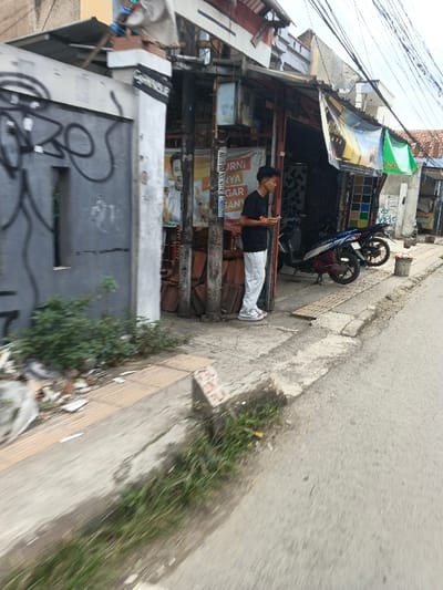 The image depicts an urban street scene in Margahayu, Indonesia, during daylight hours under an overcast sky. In the foreground, an asphalt road runs parallel to a concrete sidewalk on the left. A narrow drainage ditch separates the road from the sidewalk, overgrown with green grass and weeds.
On the sidewalk, a young male individual stands to the right of the frame's center. He wears a black short-sleeved t-shirt, light-colored (likely white) pants, and white sandals. He holds a smartphone in both hands at waist height, with his gaze directed slightly downward or to his right. His hair is dark and styled.
Behind the individual, two motorbikes are parked on the sidewalk in front of a commercial building. The closer motorbike is dark grey/black, while the second, further back, is blue and black. The building behind them features an open shopfront with an awning. Various items are visible inside, and multiple posters adorn its exterior; one depicts a smiling man, another includes the partially visible Indonesian text "MURNI HANYA SEGAR BANYA..." (purely only fresh many...). A pile of reddish-brown roof tiles or bricks is stacked near the shop entrance, and a light-blue utility box is mounted on a pole. A white plastic bucket containing fruit (apples, oranges) sits on the sidewalk further down from the motorbikes.
To the left of the male individual, a grey concrete gate or wall with black graffiti marks a property boundary. Adjacent to this gate is a white pillar bearing a dark sign with partially visible white text, "SAGAMULYAP." The sidewalk in the foreground exhibits a damaged brown tactile paving strip, and scattered debris including plastic bags and litter is present near the gate. Overhead, numerous black power lines crisscross against the sky.